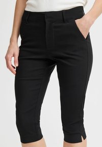 Žena koja nosi crne uske skraćene hlače s prednjim džepovima i prorezima za remen, uparene s bijelom majicom, stoji ispred jednobojne pozadine.