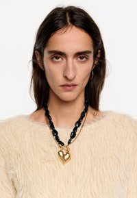 Collar de cuentas negras con un colgante en forma de corazón dorado, llevado sobre un suéter texturizado color crema, que presenta una apariencia suave y esponjosa.