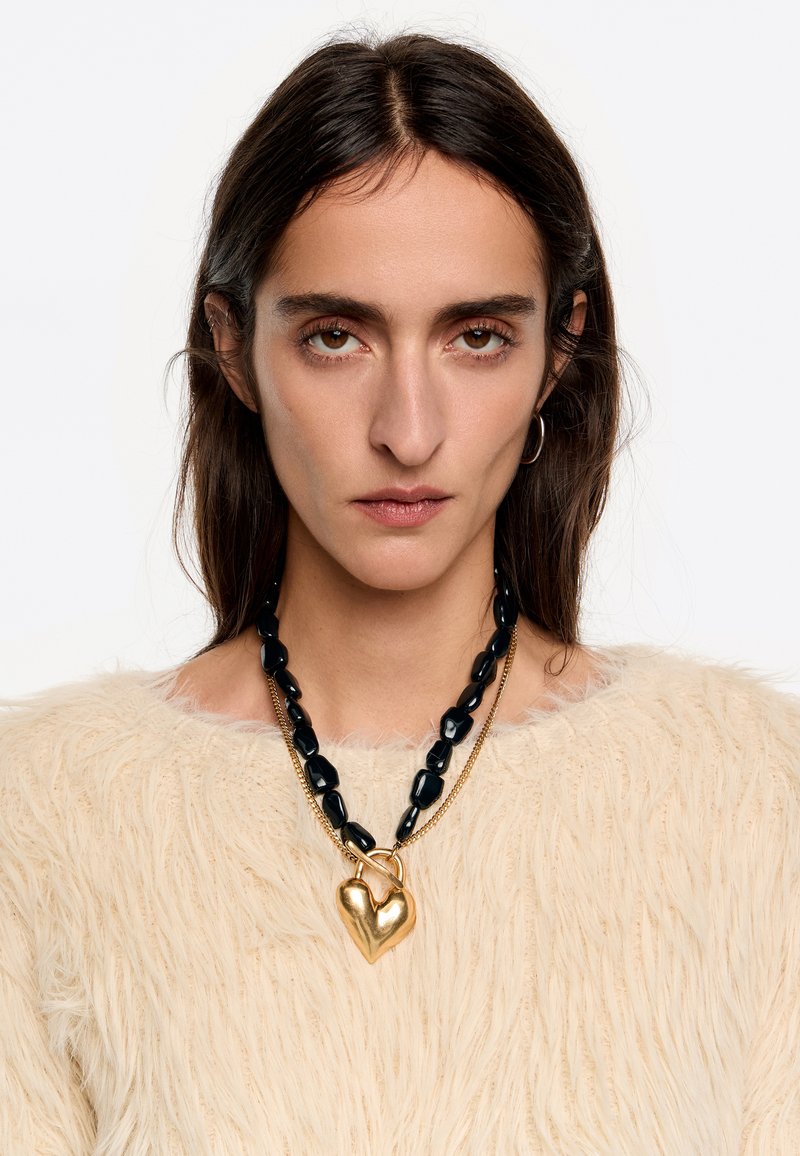 Collar de cuentas negras con un colgante en forma de corazón dorado, llevado sobre un suéter texturizado color crema, que presenta una apariencia suave y esponjosa.