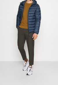 Doudoune marine avec capuche, pull texturé moutarde, pantalon marron ajusté et chaussures de sport blanches avec des accents noirs et orange.