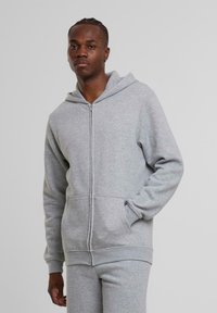 Haut à capuche gris zippé en tissu doux, avec une poche avant, un cordon de serrage à la capuche et des poignets côtelés. Porté avec un pantalon gris assorti.