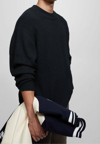 Pull en maille noire avec un col rond, présentant une texture côtelée sur les manches et le torse, porté avec une veste bleu marine et crème.