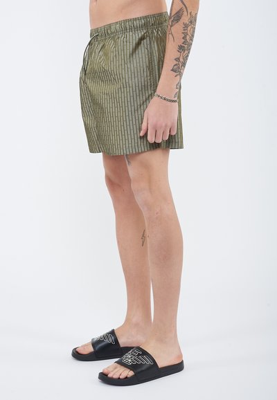 Άνδρας που φοράει ελαιίσιο πράσινο σορτς με σχέδια και μαύρα σανδάλια slide, με τατουάζ στο χέρι και στο πόδι, στέκεται μπροστά σε λευκό φόντο.