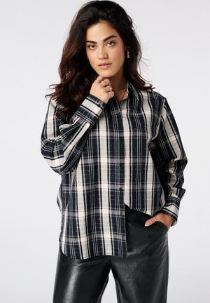 Vrouw met lang donker haar draagt een zwart-wit geruit overhemd met knopen en een zwarte leren broek, poseert met één hand in haar zak en de andere dicht bij haar gezicht.