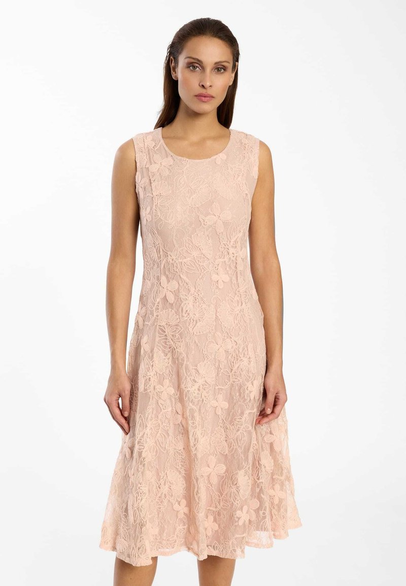 Frau trägt ärmelloses, knielanges hellrosa Spitzenkleid mit Blumenmustern, steht vor weißem Hintergrund.