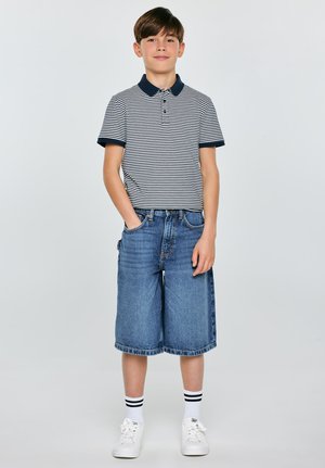 Chłopiec stojący z rękami w kieszeniach, ubrany w pasiastą koszulkę polo, dżinsowe krótkie spodenki do kolan, białe skarpety z czarnymi paskami oraz białe trampki.
