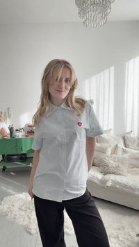 Blonde vrouw die een lichtblauw gestreept overhemd draagt met een rode hartspeld, staand in een lichte woonkamer met een bank en een groene kast achter haar.