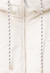 Ljuskrämfärgad zip-jacka med hög krage, svart och vit mönstrad dragsko, och ett slätt, strukturerat tyg.