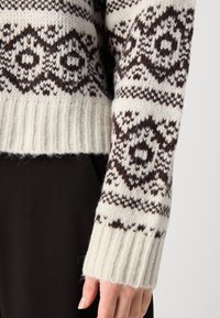 Maglione lavorato a maglia color crema con motivi geometrici marrone scuro. Presenta polsini e vita a costine, mostrando un tessuto morbido e testurizzato.
