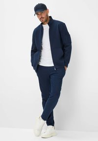Navy zip-up jacket with high collar, paired with matching pants and white sneakers. Smooth fabric with a side pocket detail on the jacket.