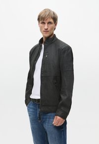 Czarna skórzana kurtka z wysokim kołnierzem, zapinana na zamek z przodu, z pikowanymi detalami na ramionach, zestawiona z białą koszulką i niebieskimi dżinsami.