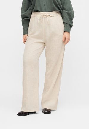 Άτομο που φοράει μπεζ παντελόνι φαρδύ στο πόδι με λάστιχο στη μέση, σκουροπράσινο μπλουζάκι με μακριά μανίκια και σκούρα παπούτσια με τετράγωνη μύτη, στέκεται σε λευκό φόντο.