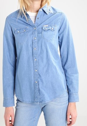 Personne portant une chemise en jean bleu clair à boutons avec deux poches poitrine, associée à un jean en denim bleu clair sur un fond uni.