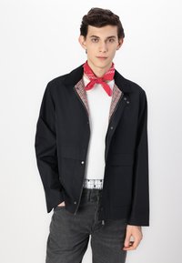 Black zip-up jacket with two front pockets, textured collar, and a red patterned scarf tied at the neck, worn over a white sweater.