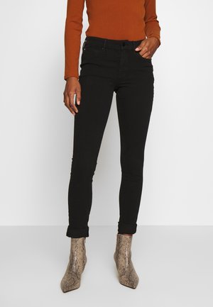 Frau trägt schwarze Skinny-Hose mit Bündchen, orangefarbenes, geripptes Langarmshirt und spitze Knöchelstiefel mit Schlangenleder-Muster.