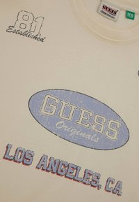 Jasno beżowa koszulka z niebieskim owalnym logo z napisem "GUESS Originals" oraz "LOS ANGELES, CA" w czerwonym i niebieskim haftem. Tkana tkanina.