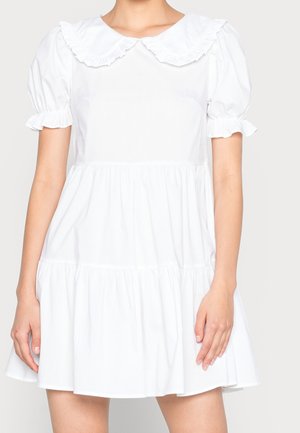 Femme portant une courte robe blanche avec des manches bouffantes, des poignets à volants et un grand col à volants, debout devant un fond uni.