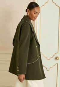 Maslinasto zelena sakoa s velikim ovratnikom, kontrastnim bijelim šavovima, prednjim džepovima i zlatnim dugmadi na rukavima.