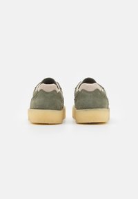 Green suede sneakers with a beige suede accent at the heel, featuring a rubber sole and rounded design, viewed from the back.