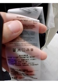 Etichetta di cura trasparente, con testo e simboli multicolori, tenuta con le dita; include istruzioni per il lavaggio, l'asciugatura e la stiratura.