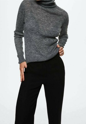 Personne portant un pull à col roulé gris ajusté et un pantalon noir taille haute, les mains posées sur les hanches.