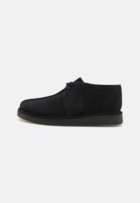 Chaussure en daim noir avec un design à bout rond, un devant à lacets et une semelle en caoutchouc texturée. Présente des coutures minimales et une finition lisse.