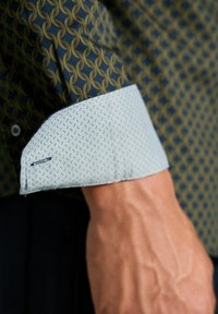 Langarmhemd mit einem dunkelblauen und goldenen geometrischen Muster, einem hellblauen gemusterten Bündchen und sichtbaren Nähedetails.
