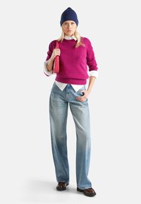 Růžový pletený svetr, bílá košile s límečkem pod ním, světle modré kalhoty s širokými nohavicemi, námořnická čepice, držící červenou kabelku, hnědé boty.