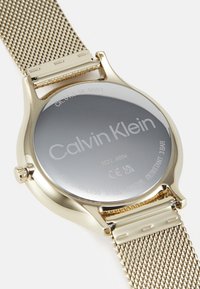 La montre en acier inoxydable Calvin Klein présente un fond argenté gravé avec des accents dorés et un bracelet en maille, mettant en avant un design épuré et moderne.