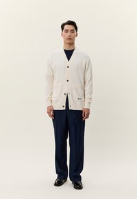 Man som står och bär en gräddfärgad kofta med knappar och framfickor, mörkblå byxor, svarta skor, mot en enkel ljus bakgrund.