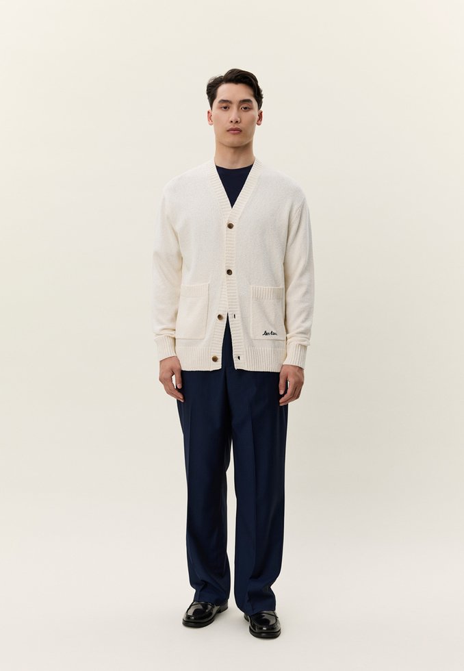 Man som står och bär en gräddfärgad kofta med knappar och framfickor, mörkblå byxor, svarta skor, mot en enkel ljus bakgrund.