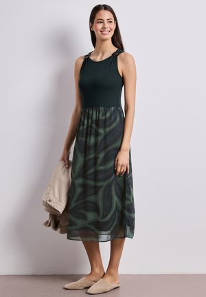 Kvinde smiler, iført en ærmeløs sort topkjole med grøn abstrakt mønstret nederdel, holder en beige jakke og har beige slip-on sko på.