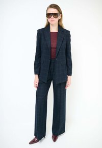 Completo a quadri blu navy con blazer strutturato e pantaloni a gamba larga, abbinato a una maglia color marsala e occhiali da sole marroni oversize. Scarpe con tacco lucide color burgundy.