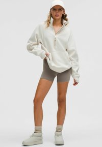 Ženska, oblečena v bež kapo, oversized svetlo kremast zip-pončo, taupe kolesarske kratke hlače, ohlapne nogavice in bež platformske čevlje, stoji na beli podlagi.