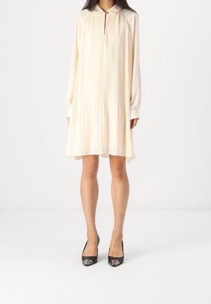 Vestido plisado color crema pálido con mangas largas, escote en forma de ojo de cerradura y detalles plateados; combinado con tacones negros con punteras metálicas.