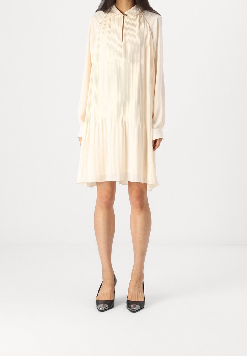 Robe plissée crème pâle avec manches longues, encolure goutte d'eau et accents argentés ; associée à des talons noirs avec bouts métalliques.