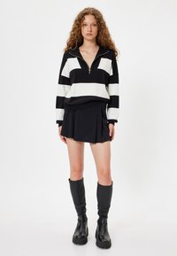 Schwarz-weißer gestreifter Pullover mit einem Reißverschlusskragen, kombiniert mit einem schwarzen plissierten Minirock und knielangen schwarzen Stiefeln. Glattes Strickmaterial.