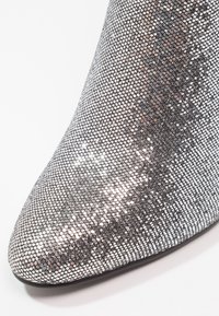 Gros plan sur l'avant d'une chaussure pointue recouverte de sequins argentés, avec une surface réfléchissante et texturée sur un fond blanc.