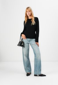 Młoda kobieta z długimi blond włosami, ubrana w czarny sweter, jasnoniebieskie spodnie typu wide-leg, czarne buty i trzymająca małą czarną torebkę na tle białym.