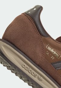 Brown suede and textile sneaker with a smooth leather heel, gold logo accents, and a textured rubber outsole featuring a zigzag pattern.
