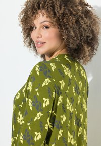 Frau mit lockigem Haar, die ein grünes Langarmoberteil mit gelben und blauen Blumenmustern trägt, sich über die Schulter umschaut und lächelt.