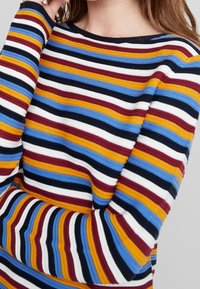 Pull en maille rayé avec des lignes horizontales de couleurs : jaune, rouge, bleu, noir et blanc. Col rond classique et manches longues. Texture douce.