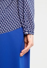 Blusa a maniche lunghe di colore blu navy con un motivo a foglie rosa, abbinata a una gonna blu uniforme. La blusa presenta polsini con bottoni e una texture liscia.