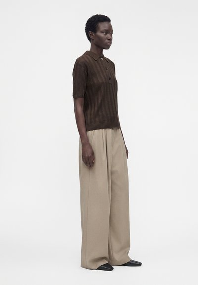 Femme aux cheveux courts et bouclés portant un polo marron à manches courtes boutonné, un pantalon large beige et des chaussures noires, debout devant un fond blanc.