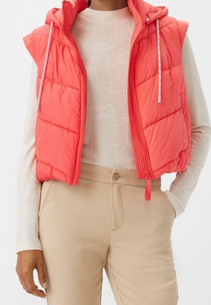 Coral quilted sleeveless puffer vest with hood worn over a white long-sleeve patterned top and beige high-waisted pants.