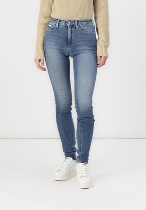 Jean skinny bleu avec taille haute, en denim. Comprend cinq poches, une fermeture à bouton et un léger dégradé. Associé à des baskets blanches.