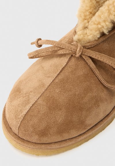 Chinelo de camurça castanho com laço no topo e forro interior de pelagem fofa, apresentando uma sola de borracha.