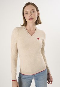 Beige V-halstrui met lange mouwen, voorzien van een kleine rode hartborduursel op de borst en een rode rand aan de onderzijde. Zachte textuur.