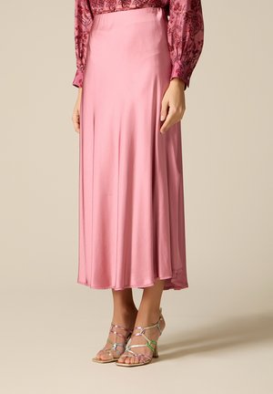 Donna che indossa una gonna midi rosa in satin fluente, abbinata a sandali con tacco multicolori e con cinturini, in piedi davanti a uno sfondo semplice.