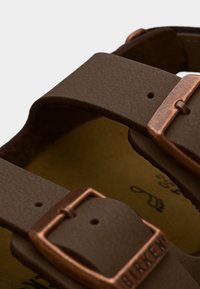 Εγγύς πλάνο καφέ δερμάτινων λουριών σανδαλιών με χάλκινες αγκράφες και ανάγλυφο το όνομα της μάρκας στο κάτω λουρί.